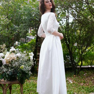 May include: A white midi dress with lace sleeves and a round neckline. The dress is worn outdoors in a garden setting. The model is wearing nude-colored heels. A bouquet of white flowers sits on a wooden table.