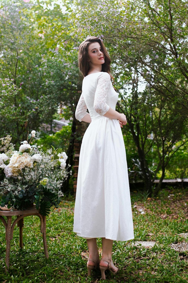 May include: A white midi dress with lace sleeves and a round neckline. The dress is worn outdoors in a garden setting. The model is wearing nude-colored heels. A bouquet of white flowers sits on a wooden table.