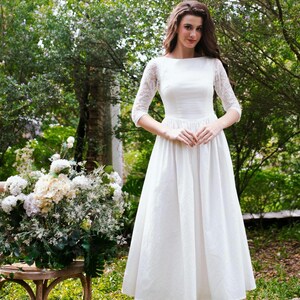 May include: A white midi dress with lace sleeves and a boat neckline. The dress has a fitted bodice and a flared skirt. The person is standing in a garden setting with greenery and flowers.