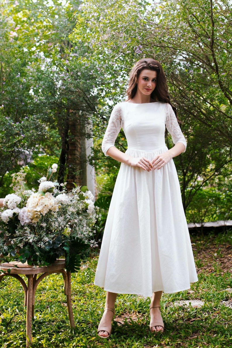 May include: A white midi dress with lace sleeves and a boat neckline. The dress has a fitted bodice and a flared skirt. The person is standing in a garden setting with greenery and flowers.