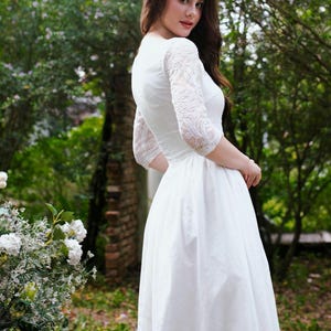 May include: A white wedding dress with lace sleeves. The dress has a fitted bodice and a flowing skirt. The sleeves are three-quarter length and made of delicate lace. The dress is worn outdoors in a garden setting.