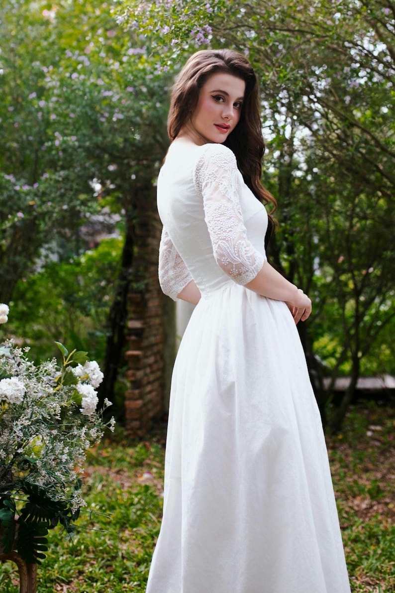 May include: A white wedding dress with lace sleeves. The dress has a fitted bodice and a flowing skirt. The sleeves are three-quarter length and made of delicate lace. The dress is worn outdoors in a garden setting.