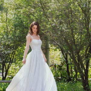 May include: A woman wearing a white, flowing dress stands in a grassy area with trees in the background. The dress has thin straps and a sweetheart neckline.