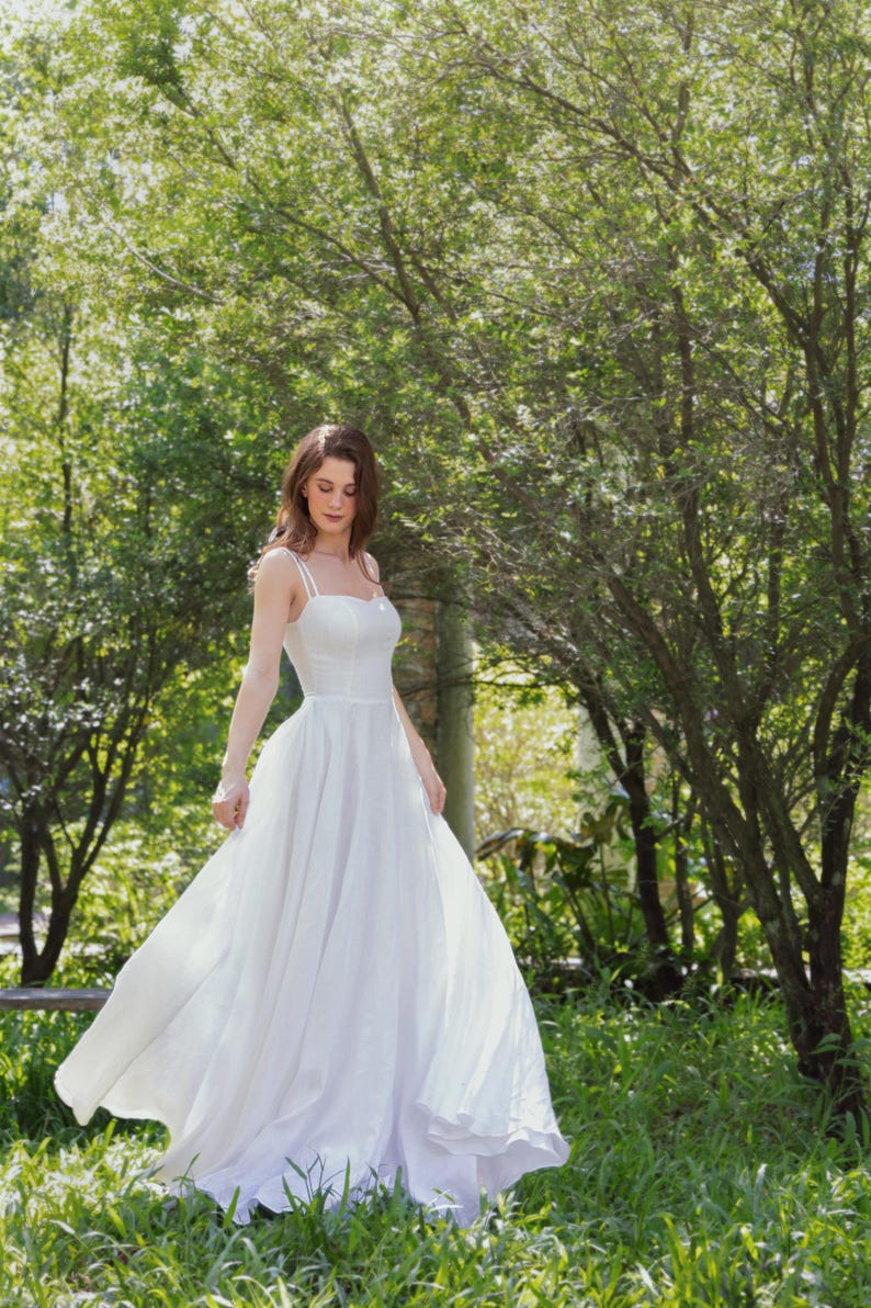 May include: A woman wearing a white, flowing dress stands in a grassy area with trees in the background. The dress has thin straps and a sweetheart neckline.
