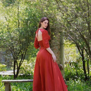May include: A full-length, red dress with a low back and three-quarter sleeves. The dress is made of a light fabric and has a full skirt. The model is in a garden with trees and greenery, creating a natural backdrop.