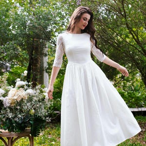 May include: A white wedding dress with a boat neckline and a fitted bodice. The dress features lace sleeves and a flowing skirt. The image is set outdoors with a bouquet of flowers on a wooden table.