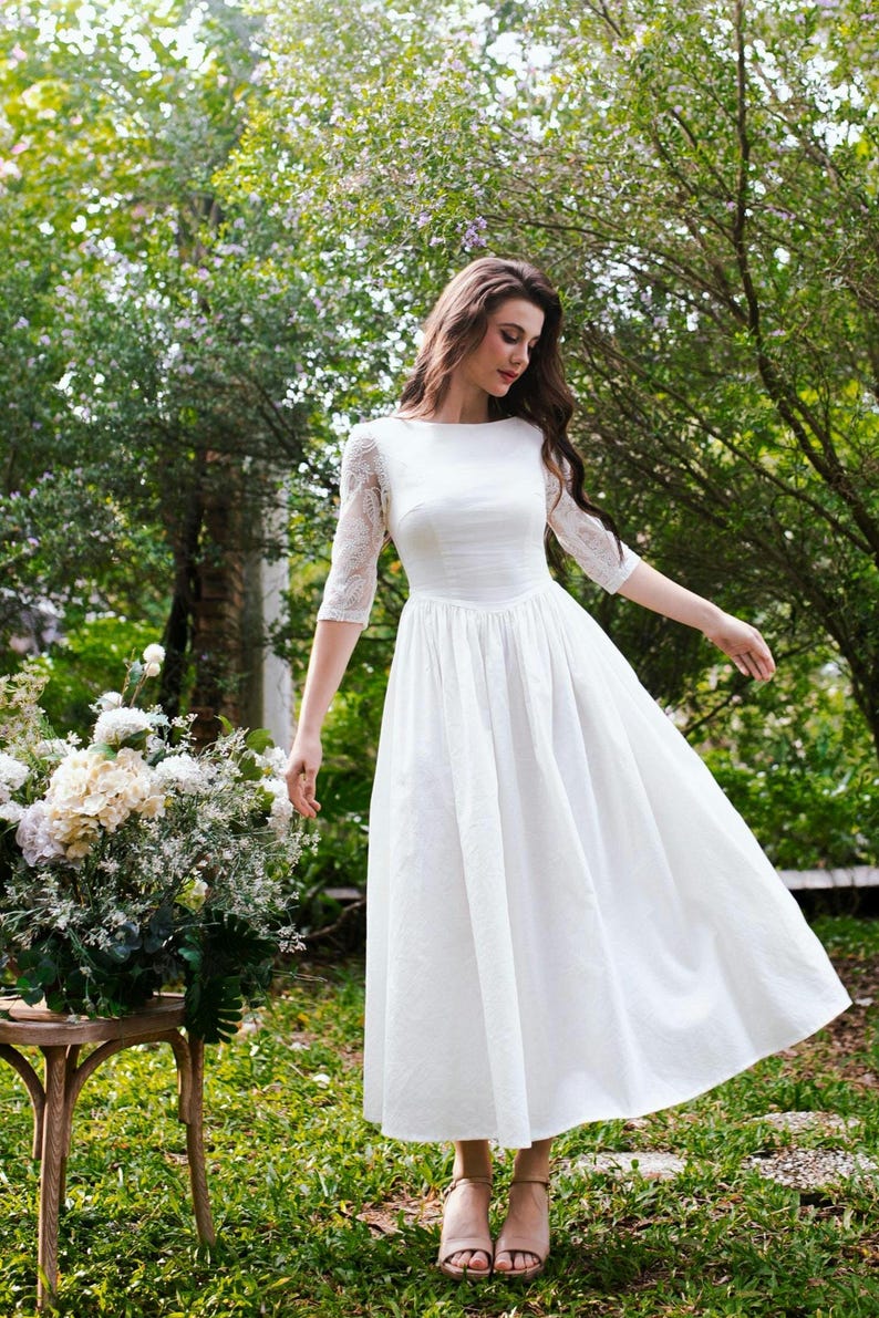 May include: A white wedding dress with a boat neckline and a fitted bodice. The dress features lace sleeves and a flowing skirt. The image is set outdoors with a bouquet of flowers on a wooden table.