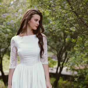 May include: A white wedding dress with a boat neckline and three-quarter sleeves. The sleeves are made of lace. The dress has a fitted bodice and a full skirt. The background is a lush green garden with trees and foliage.