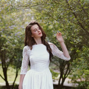 May include: A white wedding dress with a boat neckline and three-quarter sleeves with lace detailing. The dress has a fitted bodice and a flowing skirt. The background features lush greenery and trees, creating a natural and romantic setting.