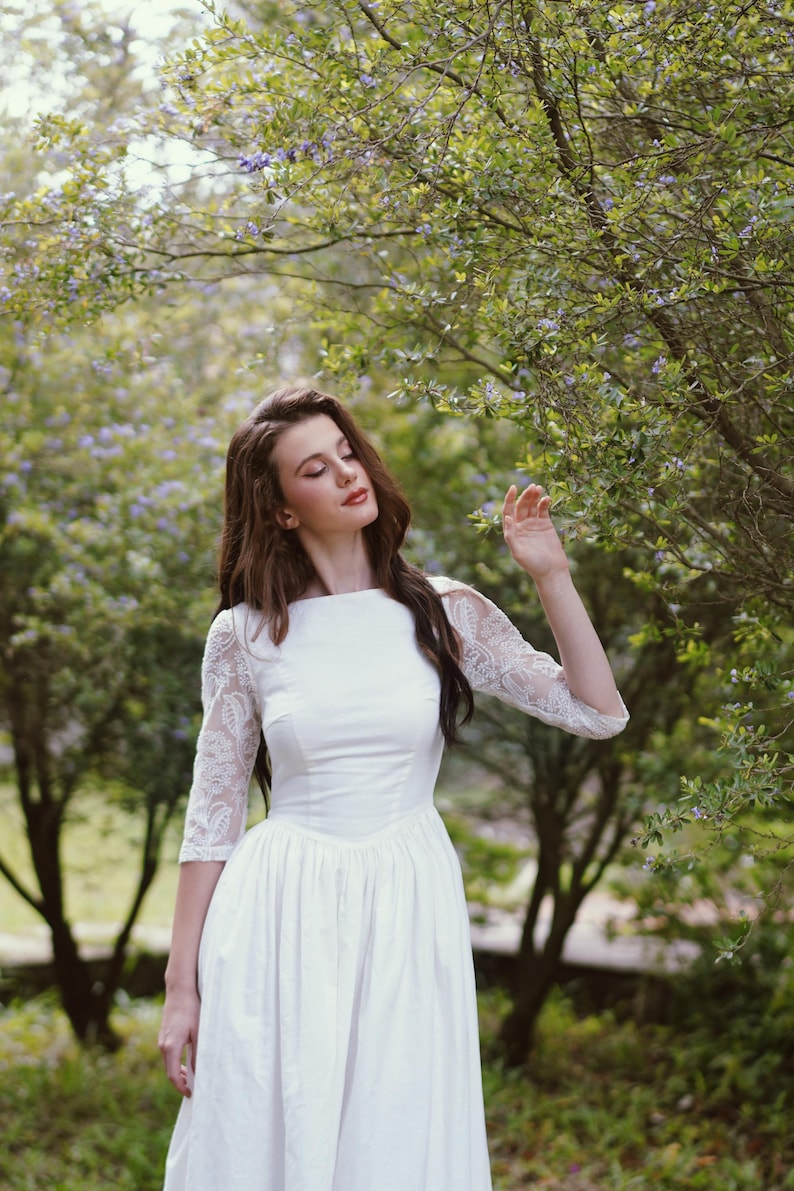 May include: A white wedding dress with a boat neckline and three-quarter sleeves with lace detailing. The dress has a fitted bodice and a flowing skirt. The background features lush greenery and trees, creating a natural and romantic setting.