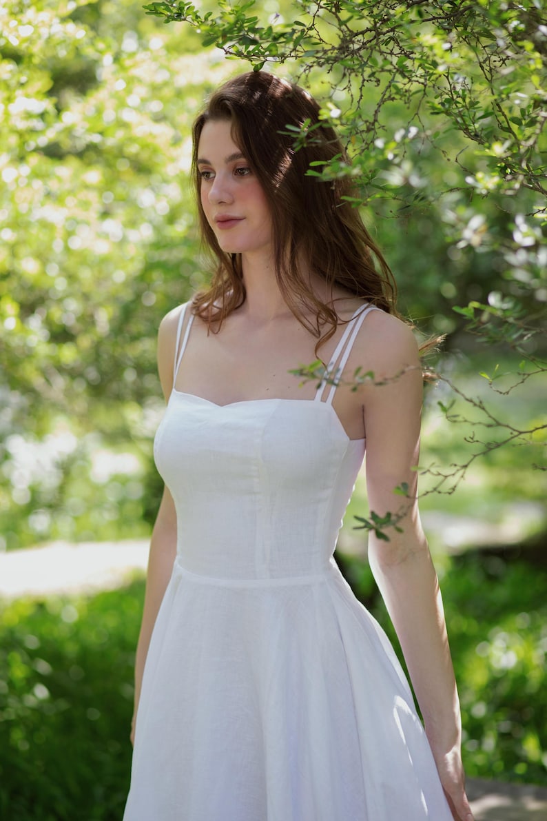 May include: A woman wearing a white linen sundress with thin straps. She is standing in a garden with green foliage and sunlight.