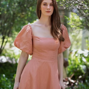 May include: A peach-colored dress with a square neckline and short, puffed sleeves. The dress has a fitted bodice and a flared skirt. The model is standing outdoors in front of a blurred green background.