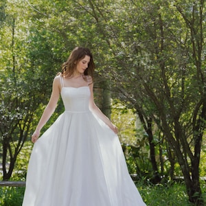 May include: A woman in a white wedding dress stands in a grassy area with trees in the background. She is holding the skirt of her dress with one hand and looking down.