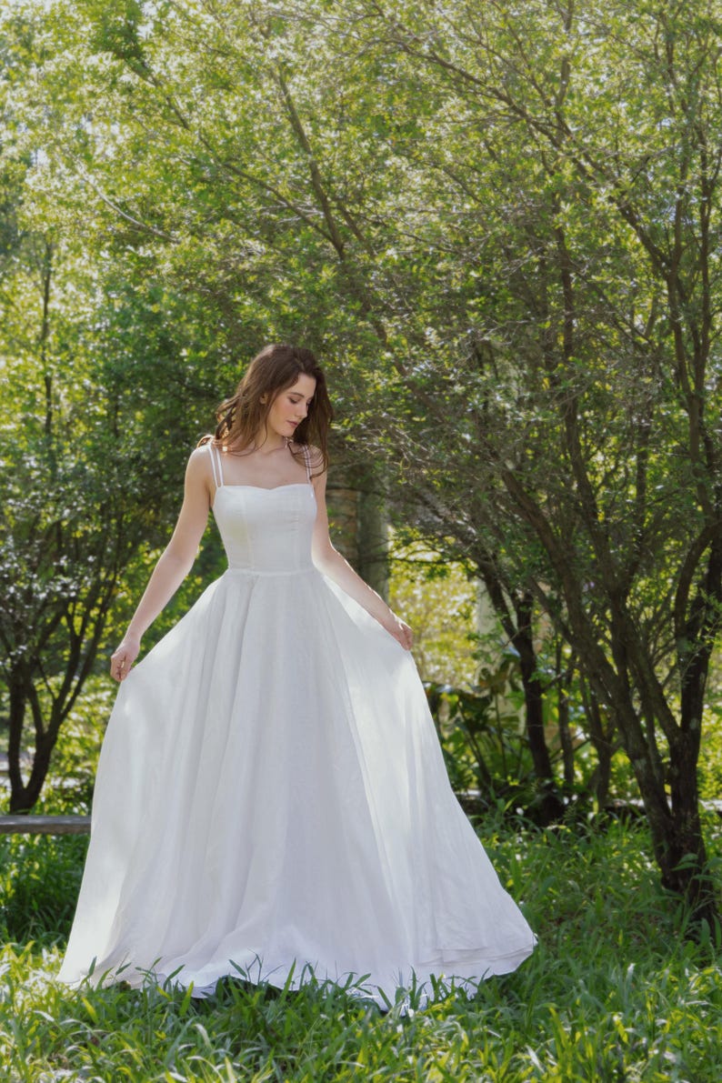 May include: A woman in a white wedding dress stands in a grassy area with trees in the background. She is holding the skirt of her dress with one hand and looking down.