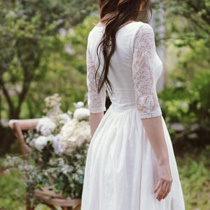 May include: A white wedding dress with three-quarter sleeves featuring intricate beaded lace details. The dress has a fitted bodice and a flowing skirt. The image is taken outdoors, with a blurred background of greenery and a wooden chair.