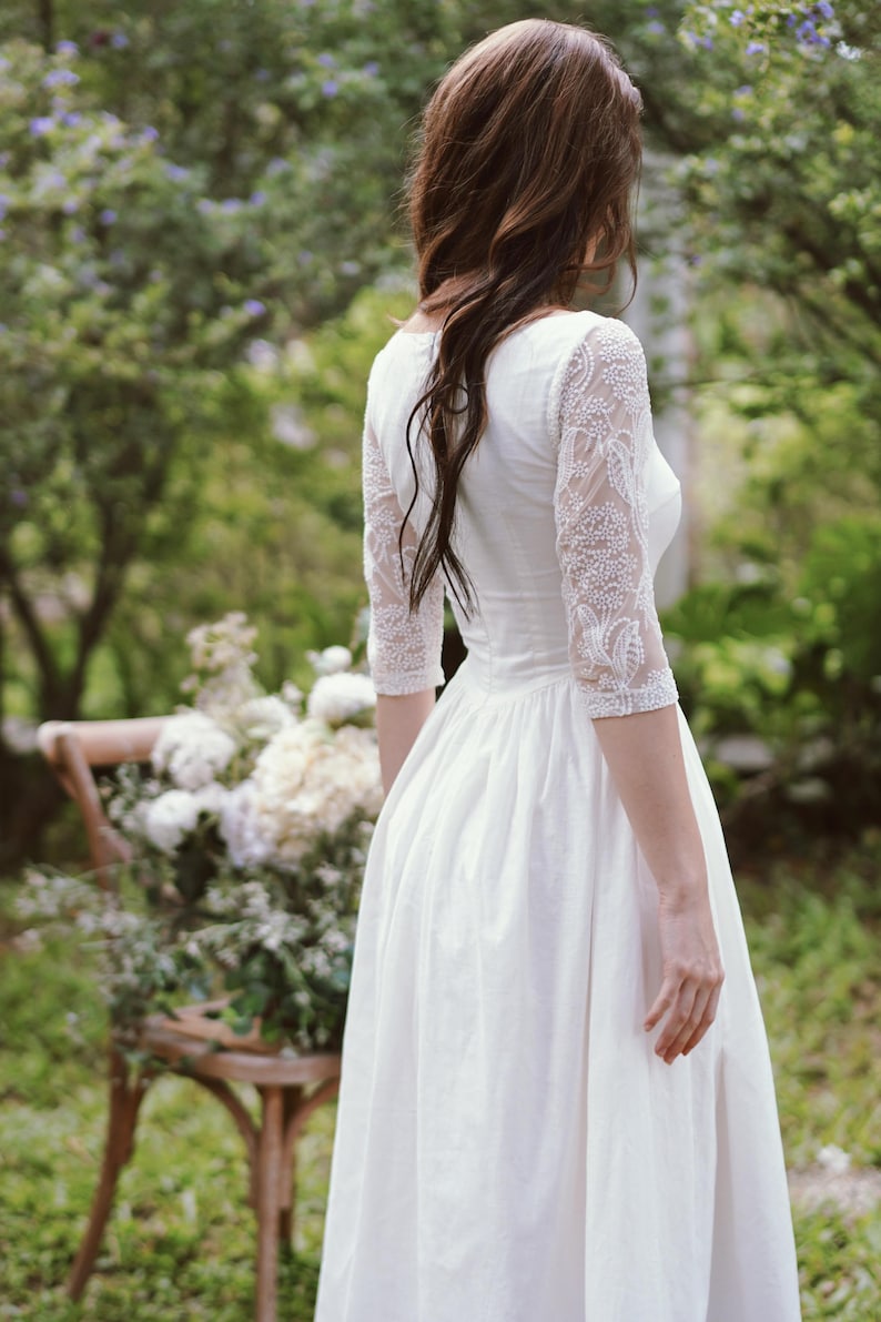 May include: A white wedding dress with three-quarter sleeves featuring intricate beaded lace details. The dress has a fitted bodice and a flowing skirt. The image is taken outdoors, with a blurred background of greenery and a wooden chair.