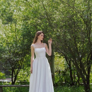 May include: A woman wearing a white sleeveless dress stands in a grassy area with trees in the background. She is looking to the right and her hand is raised.