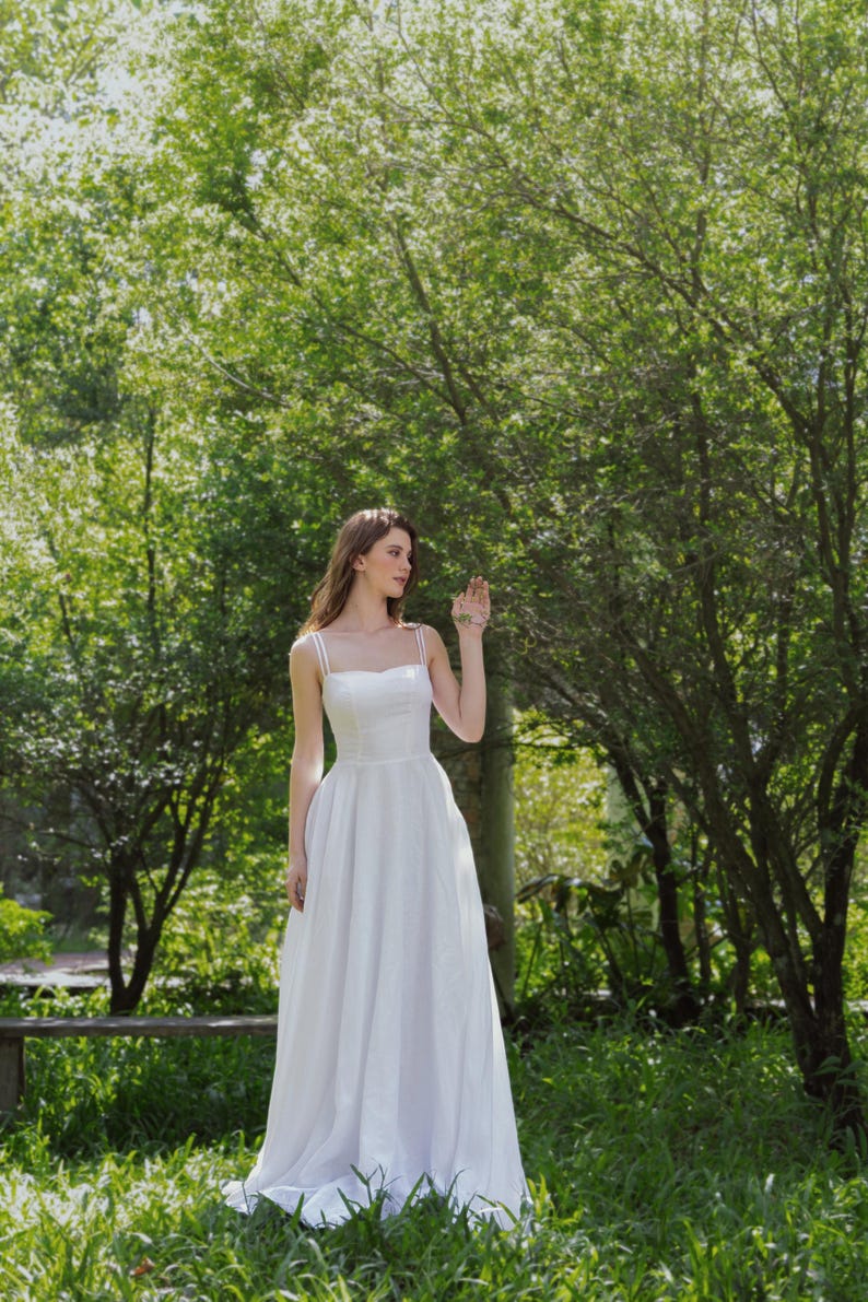 May include: A woman wearing a white sleeveless dress stands in a grassy area with trees in the background. She is looking to the right and her hand is raised.