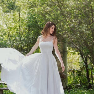 May include: A woman wearing a white, flowing dress stands in a grassy area with trees in the background. The dress has a simple, strapless design and a flowing skirt.