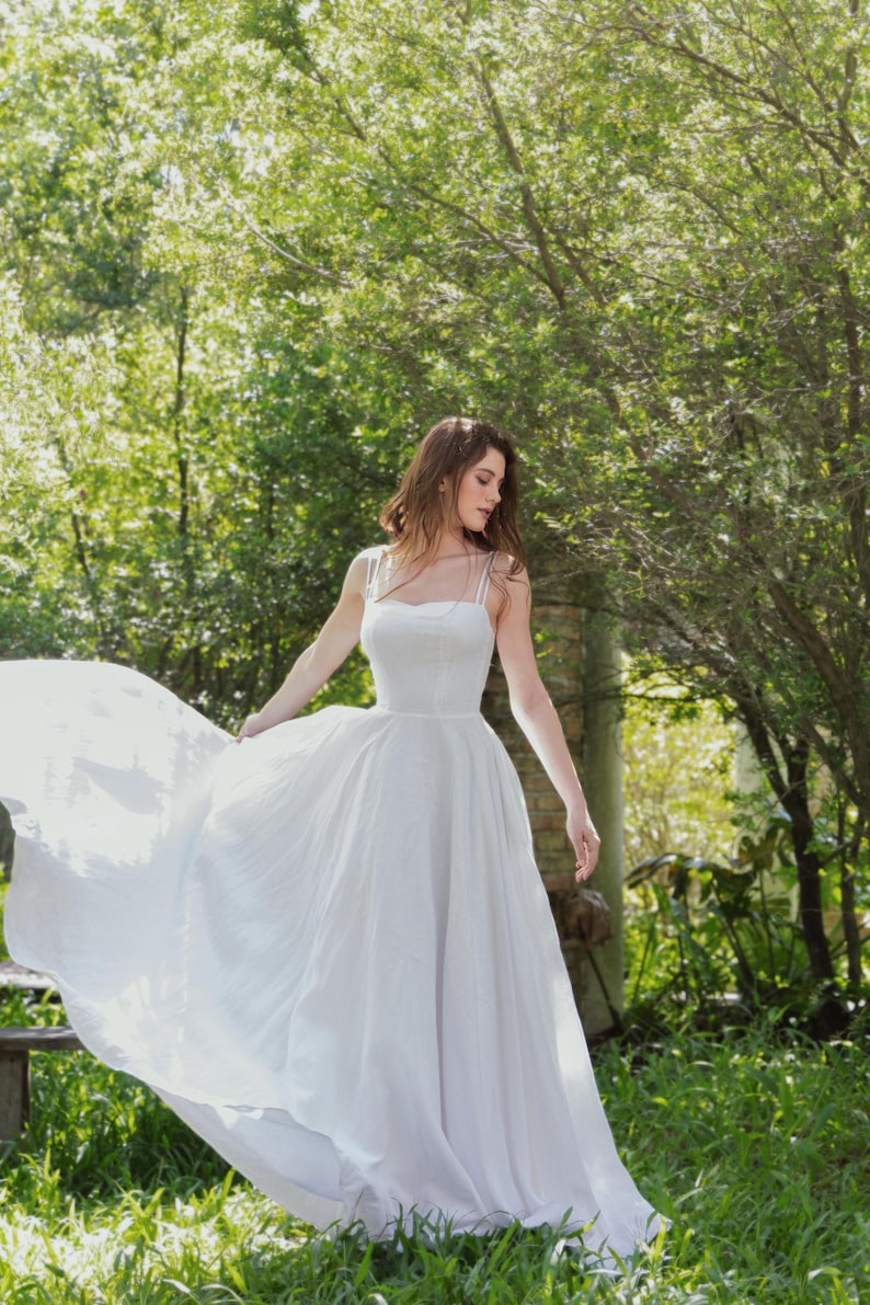 May include: A woman wearing a white, flowing dress stands in a grassy area with trees in the background. The dress has a simple, strapless design and a flowing skirt.