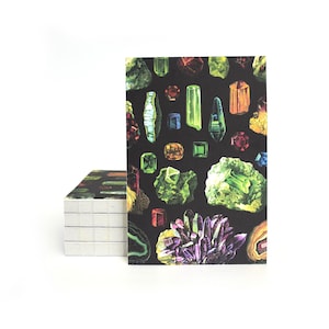May include: A stack of notebooks with a black cover featuring a colorful illustration of various gemstones. The gemstones are in shades of green, red, yellow, and purple against a black background. The notebooks are on a white background.