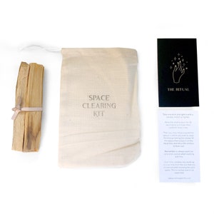 May include: A white cloth bag with the words "Space Clearing Kit" printed on it. The bag contains two bundles of dried sage sticks. A black card with the words "The Ritual" printed on it is next to the bag. The card includes instructions for using the sage sticks to clear negative energy from a space.