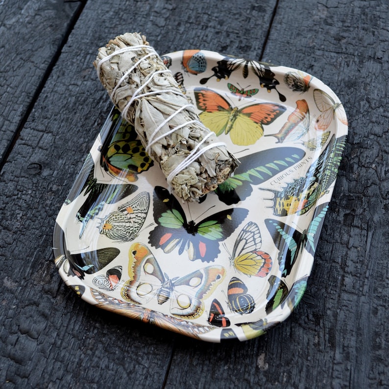 May include: A white metal tray with a butterfly design and the text "Curious Prints". A white sage smudge stick is on top of the tray.
