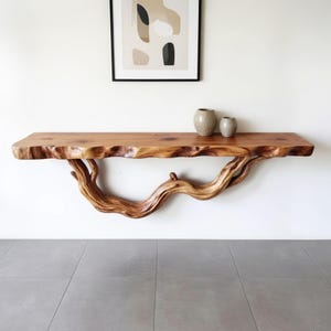 May include: A natural wood floating shelf with a live edge and a branch-like support. The shelf is a warm brown color and has two small, neutral-toned vases on top. The shelf is mounted on a white wall, creating a rustic and organic aesthetic.