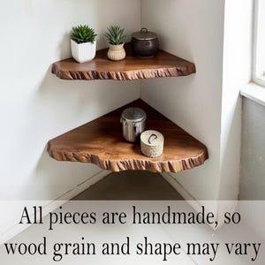 Op de afbeelding: Twee handgemaakte, driehoekige houten hoekplanken met levende randen zijn in een hoek gemonteerd. De planken zijn gebeitst in een rijke bruine kleur en bevatten kleine potplanten en decoratieve containers. De tekst onderaan luidt: "All pieces are handmade, so wood grain and shape may vary."