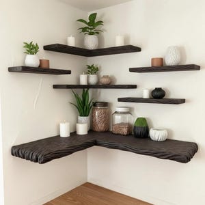 Puede incluir: Estanterías de esquina de madera marrón oscuro con una superficie texturizada, dispuestas en una esquina. Las estanterías están decoradas con pequeñas plantas en macetas, velas y jarrones decorativos. Las estanterías están montadas en una pared blanca.