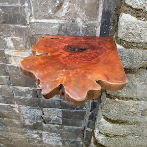 Puede incluir: Estantería de madera con borde vivo, de forma natural y orgánica. La estantería es de color marrón rojizo cálido con patrones de vetas oscuros e intrincados. Está montada en una pared de piedra gris, mostrando su diseño rústico único. Profundidad aproximada: 30 cm.