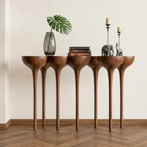 Puede incluir: Una consola de madera moderna con un diseño único. La mesa presenta una serie de superficies en forma de cuenco sostenidas por patas delgadas y alargadas. Está adornada con objetos decorativos, como un jarrón con una hoja grande, libros y pequeñas figuras de animales. La mesa mide aproximadamente 183 cm de largo.