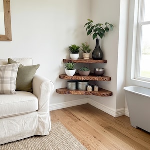 Op de afbeelding: Drie houten hoekplanken met natuurlijke randdetails, met potplanten, decoratieve potten en een donkere vaas. De planken zijn aan een witte muur bevestigd, waardoor een natuurlijke en rustieke uitstraling ontstaat. De kamer heeft een houten vloer en een witte bank.
