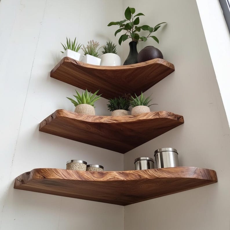 Op de afbeelding: Drie houten hoekplanken met een natuurlijke rand zijn aan een witte muur bevestigd. De planken bevatten kleine potplanten, een zwarte vaas en metalen blikken. De planken zijn gemaakt van donkerbruin hout.