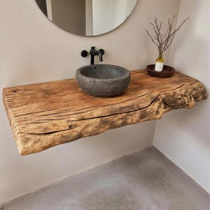 May include: A rustic bathroom vanity with a live edge wooden shelf. A gray stone vessel sink sits on the shelf, with a black faucet. A small bottle and decorative branches are on the shelf. The vanity is mounted on a light-colored wall.