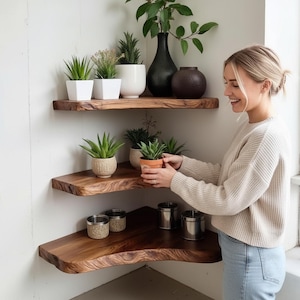 Op de afbeelding: Drie houten hoekplanken met natuurlijke randen zijn aan een witte muur bevestigd. De planken tonen diverse potplanten, waaronder vetplanten en kleine bomen, samen met decoratieve vazen en metalen containers. Een vrouw houdt een kleine potplant vast.