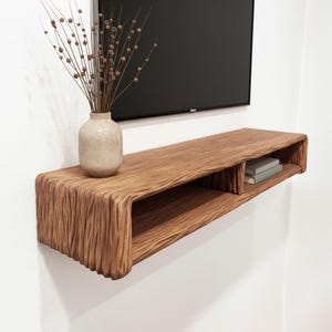 May include: A floating wooden shelf with a textured, organic design. The shelf is mounted on a white wall and features a built-in compartment. A beige vase with dried stems sits on the shelf. Two books are placed in the compartment.