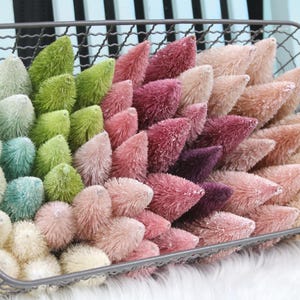 May include: A metal wire basket filled with a variety of small, colorful bottle brush trees. The trees range in color from white, teal, and green to pink, burgundy, and peach. The arrangement sits on a white, fluffy surface.