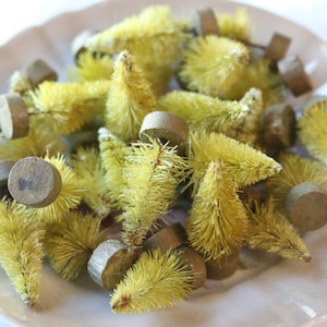 May include: A collection of small, yellow bottle brush trees with wooden bases, arranged on a white, scalloped-edge plate. The trees have a fuzzy texture and are a festive decoration.