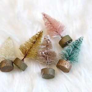 May include: A collection of small, decorative bottle brush Christmas trees in various colors, including pink, gold, and teal. Each tree is mounted on a small, round wooden base, creating a festive and charming display. The trees are arranged on a white, fluffy surface.