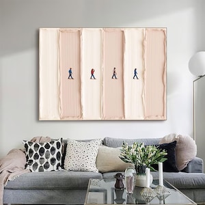 May include: Abstract wall art featuring four tiny figures walking across a textured, vertical, cream and blush-toned background. The artwork is framed in a simple, dark frame and hangs above a gray sofa with decorative pillows.