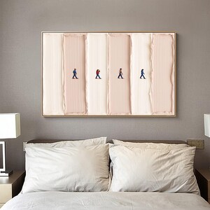 May include: A framed abstract art piece featuring four tiny figures walking across a textured, vertical, cream and blush-colored background. The artwork is encased in a gold-colored frame and hangs above a bed with white pillows.