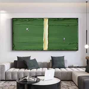 Puede incluir: Pintura abstracta de una cancha de tenis con una superficie verde texturizada y una l&iacute;nea central amarilla. Dos peque&ntilde;as figuras en blanco juegan al tenis. La obra est&aacute; enmarcada en negro y cuelga sobre un sof&aacute; gris.