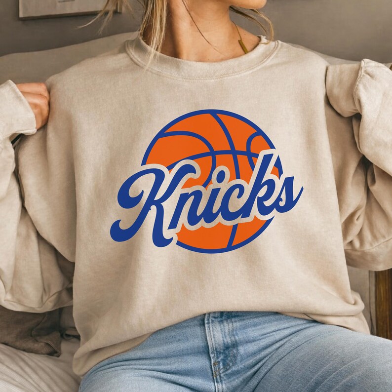 May include: Beige crew neck sweatshirt with a basketball graphic and the word "Knicks" in blue script. The basketball is orange with blue lines. The sweatshirt is a neutral colour.
