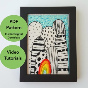 Op de afbeelding: Geborduurd kunstwerk in een zwarte lijst. Het ontwerp toont abstracte vormen in zwart en wit, met een kleurrijke regenboog aan de onderkant. De achtergrond is een lichtblauwe, getextureerde stof. De afbeelding bevat de tekst "PDF Pattern Instant Digital Download" en "Video Tutorials".