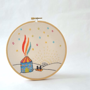 May include: Embroidered artwork in a wooden hoop. The design features a colorful building with rainbow smoke, a rising sun, and two figures on a blanket. The sky is dotted with colorful diamond shapes.