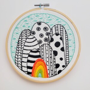 May include: Embroidered artwork in a wooden hoop. The design features stylized black and white shapes with various patterns, including stripes, dots, and geometric designs. A colorful rainbow is at the bottom, with a light blue background and a turquoise border.
