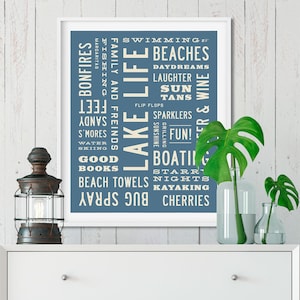 May include: A white framed print with a blue background and white text. The text is a word cloud of activities and things associated with lake life, such as swimming, beaches, bonfires, fishing, and kayaking.