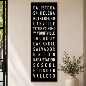 Può includere: Un cartello in legno nero, alto e rettangolare, con testo bianco in maiuscolo che elenca le località: CALISTOGA, ST. HELENA, RUTHERFORD, OAKVILLE, VETERAN'S HOME, VIA YOUNTVILLE, TRUBODY, OAK KNOLL, SALVADOR, UNION, NAPA STATION, SOSCOL, FLOSDEN, VALLEJO.