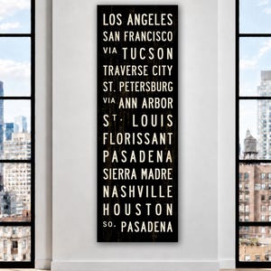 May include: A tall, rectangular black wooden sign featuring white, sans-serif text listing the cities: Los Angeles, San Francisco, via Tucson, Traverse City, St. Petersburg, via Ann Arbor, St. Louis, Florissant, Pasadena, Sierra Madre, Nashville, Houston, So. Pasadena.